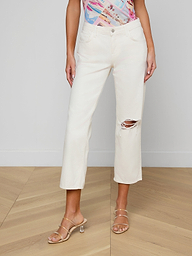A woman wears the Blaine Cropped Stovepipe Jean in cream-colored, all-cotton denim featuring a rip at one knee, paired with a colorful patterned top and nude heeled sandals, standing against a plain white background.