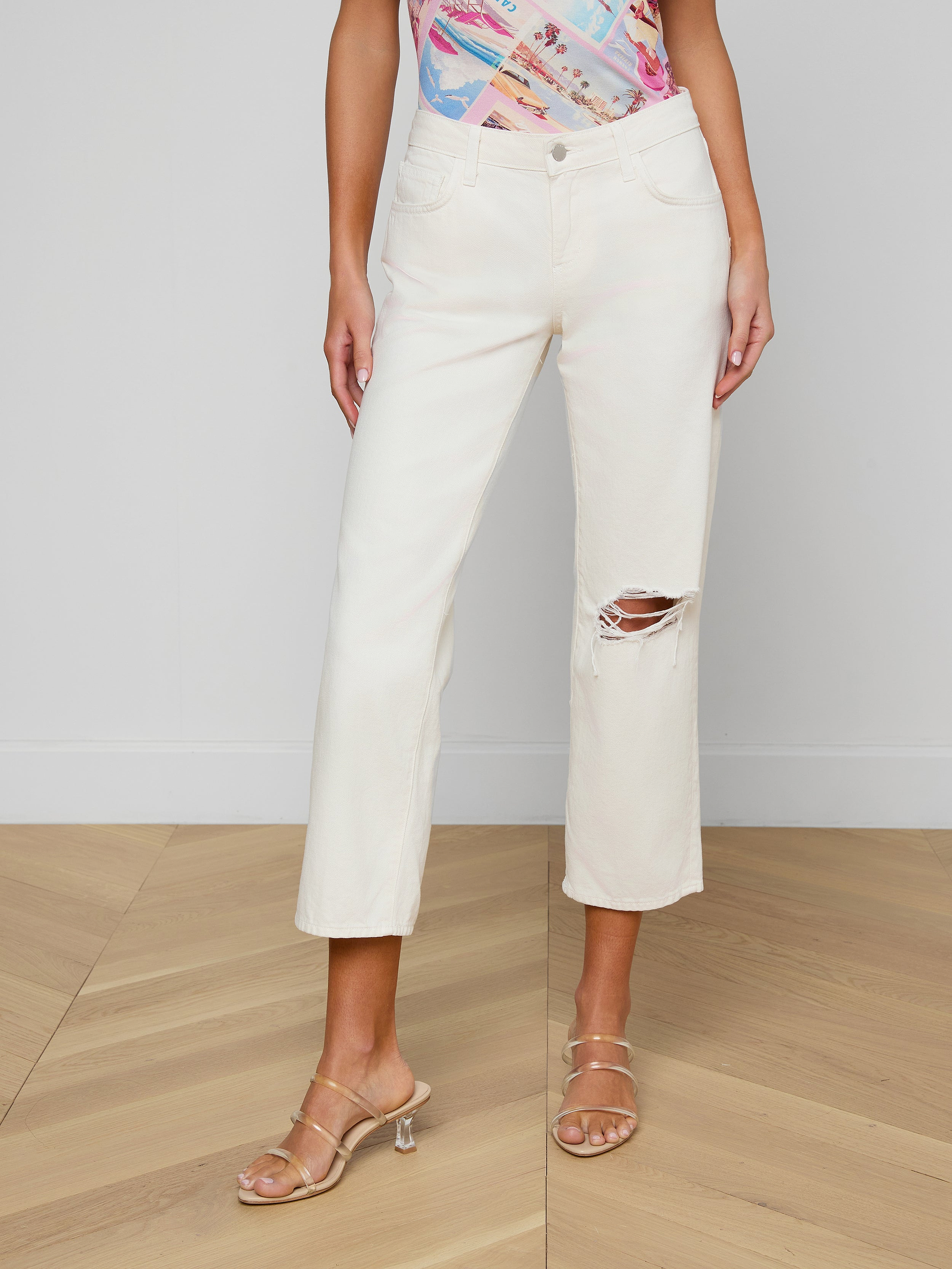 A woman wears the Blaine Cropped Stovepipe Jean in cream-colored, all-cotton denim featuring a rip at one knee, paired with a colorful patterned top and nude heeled sandals, standing against a plain white background.