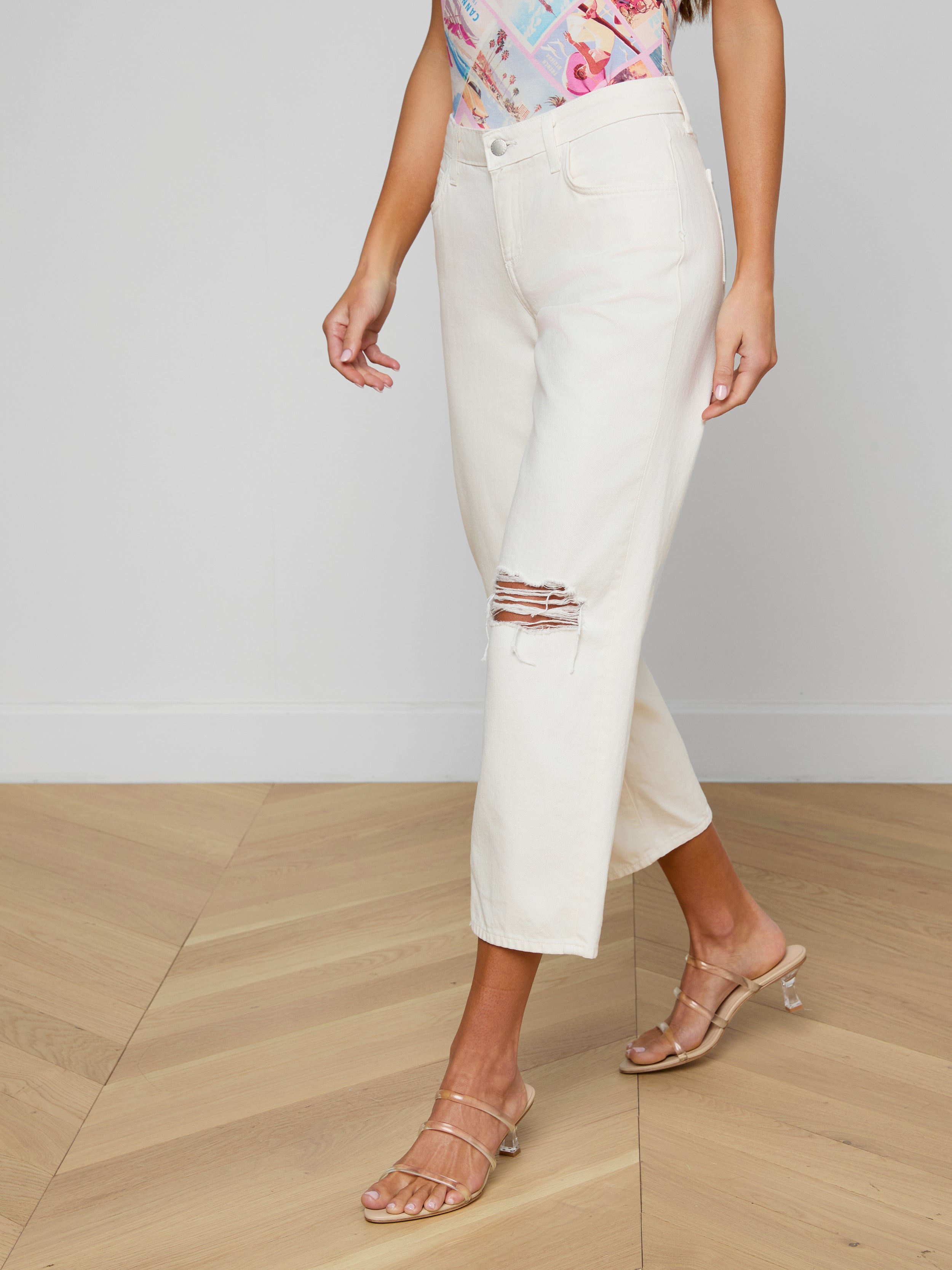 A woman wearing the Blaine Cropped Stovepipe Jean—light-colored, all-cotton denim with a ripped knee—paired with a colorful top and beige heeled sandals, stands against a plain background.