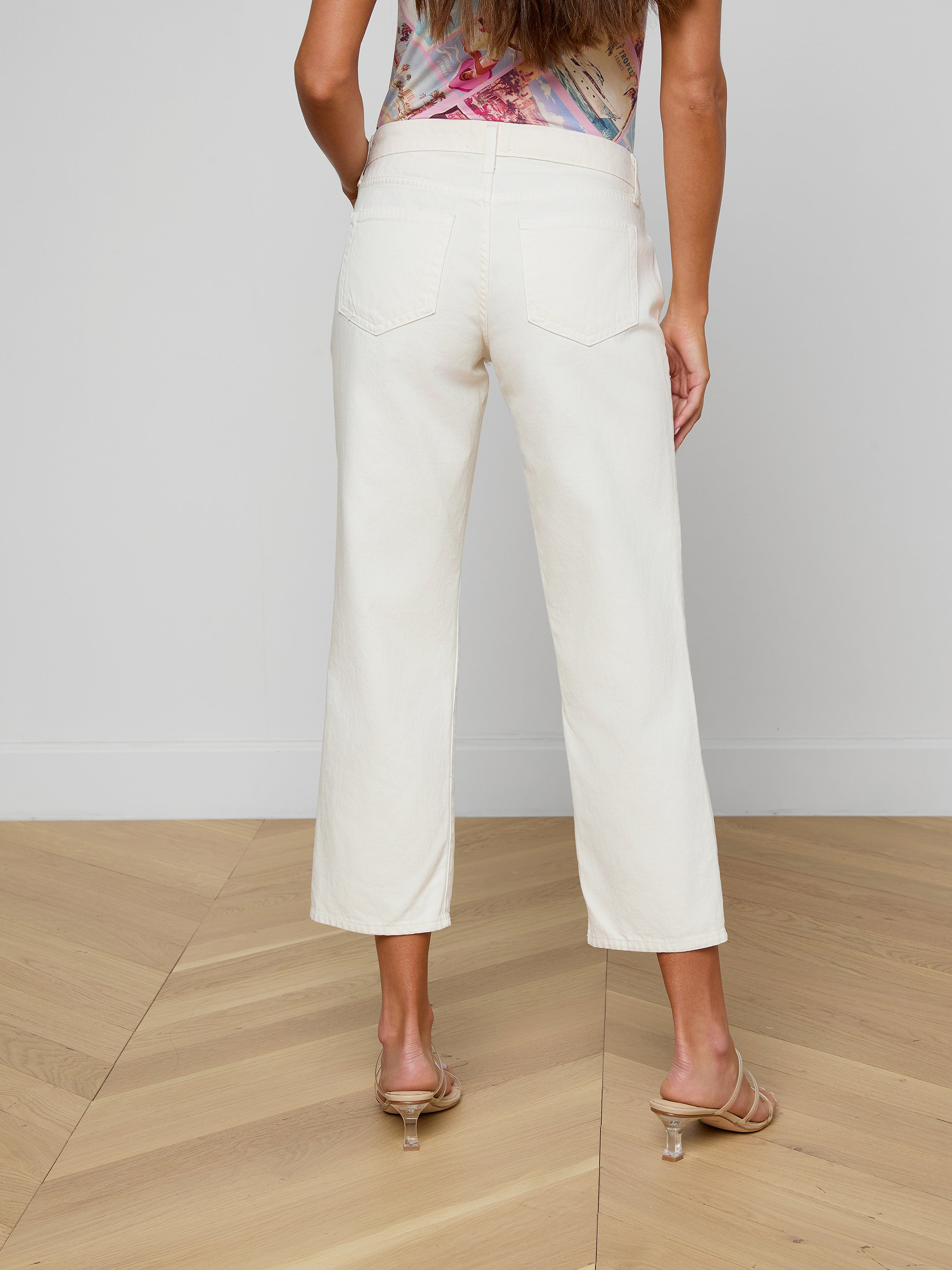 A woman is shown from the back against a plain white background, wearing the Blaine Cropped Stovepipe Jean in off-white all-cotton denim with a slouchy straight-leg fit, paired with heeled sandals.