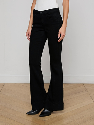 A person stands on a wooden floor against a white wall, wearing L'AGENCE Lenora Low-Rise Flare Jeans in black wash and black pointed-toe shoes. The upper body is cropped out of the image.