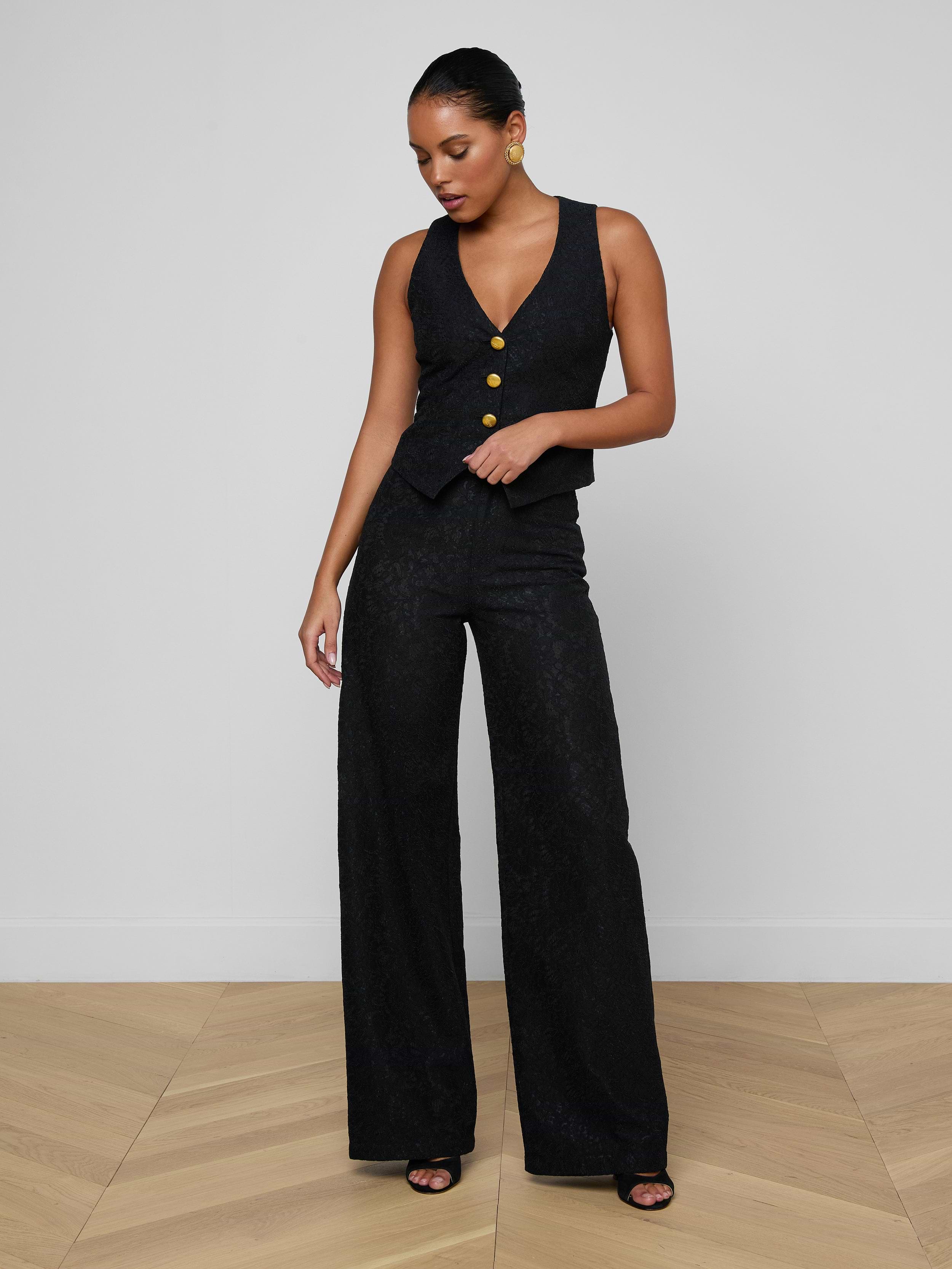 A woman stands against a plain white background wearing the Livvy Lace Trouser—a pair of high rise, wide-leg black pants—paired with a sleeveless black jumpsuit top featuring yellow buttons, black heels, and large round earrings. She looks down and rests one hand on her hip.