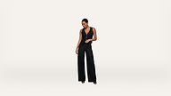 A woman stands against a plain white background wearing a black sleeveless jumpsuit and black open-toed heels, looking down with one hand in her pocket.