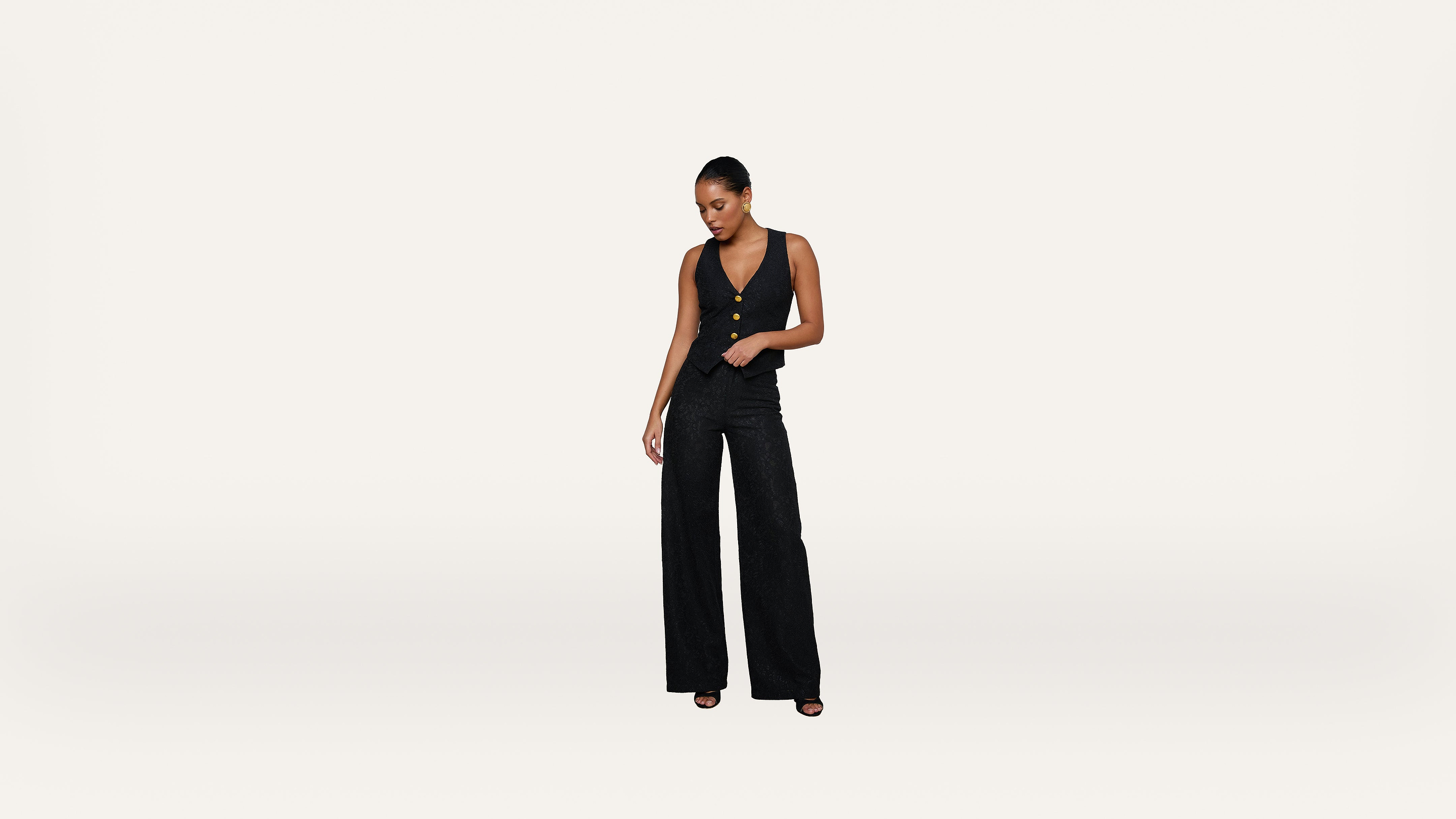 A woman stands against a plain white background wearing a black sleeveless jumpsuit and black open-toed heels, looking down with one hand in her pocket.
