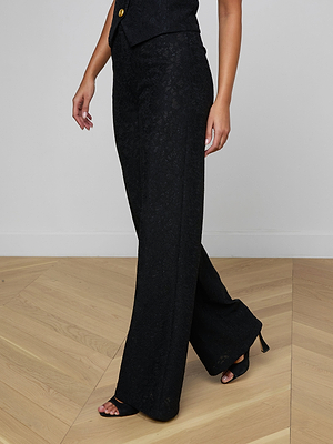 A person wearing the Livvy Lace Trouser in black with floral lace and black open-toe heels walks on a light wooden floor. Their upper body is mostly out of frame, and their left arm is slightly bent.