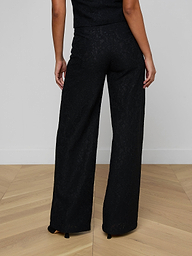 A person stands facing away, shown from the waist down against a plain light background, wearing the Livvy Lace Trouser—high-rise black floral lace trousers paired with a black top.