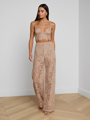 A woman with long brown hair stands indoors on a wooden floor, wearing the Livvy Lace Trouser, a cappuccino lace bralette, open-toe heels, and accessorized with earrings and a necklace.