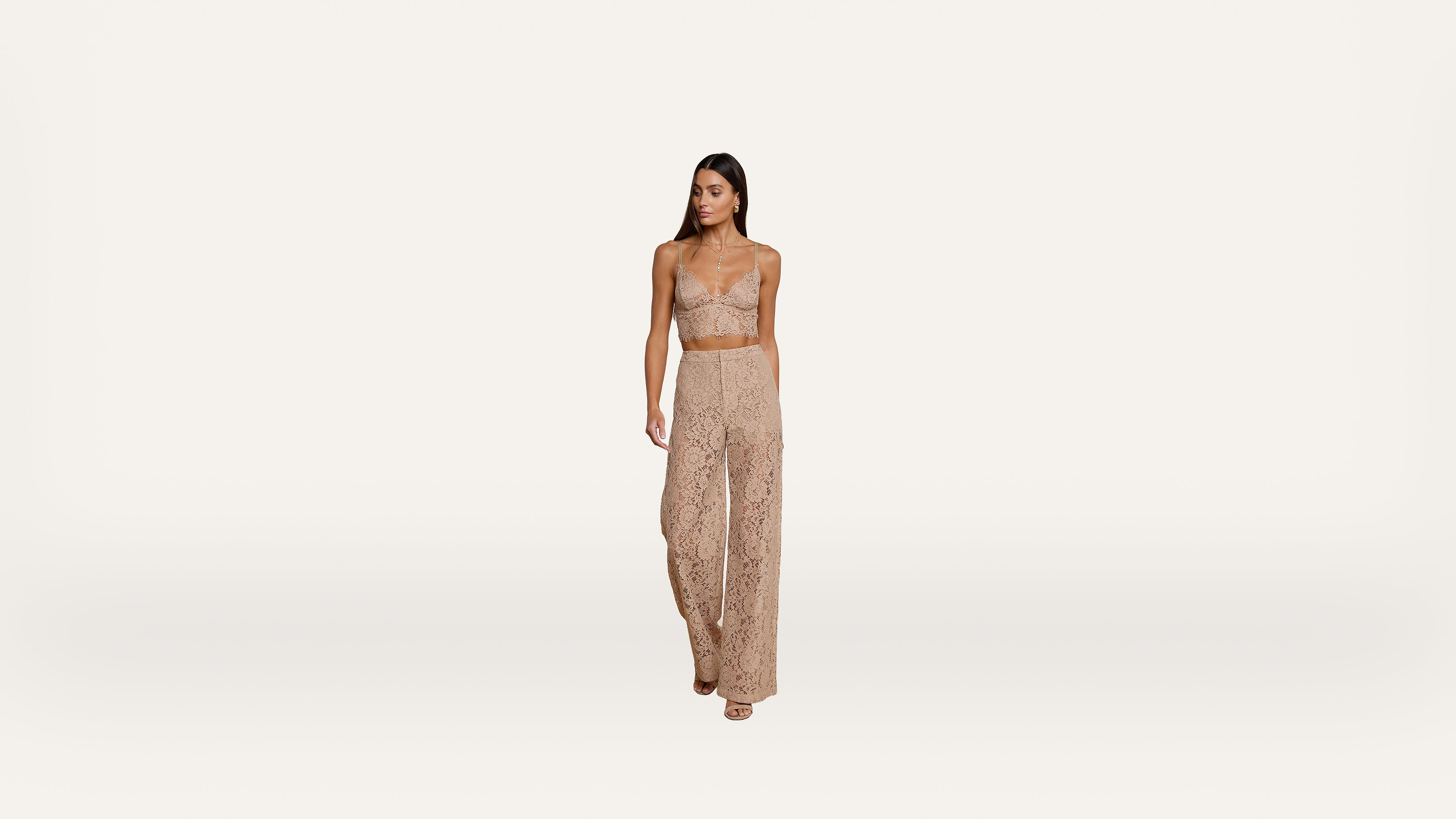 A woman in a beige lace crop top and matching high-waisted pants stands against a plain white background.