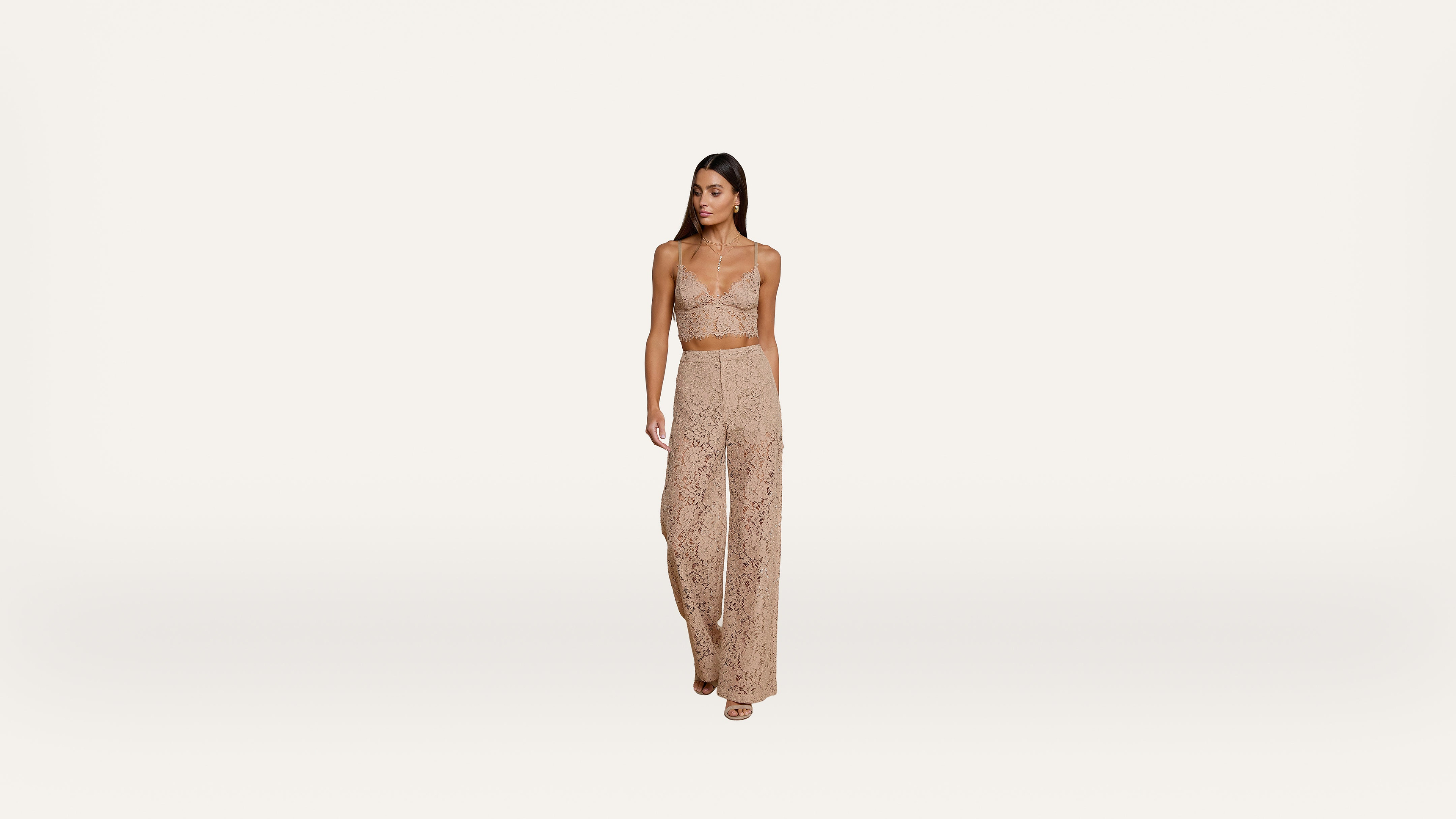A woman in a beige lace crop top and matching high-waisted pants stands against a plain white background.