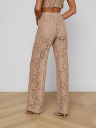 A woman is shown from the back against a plain white background, wearing the Livvy Lace Trouser—a pair of high-rise, wide-leg, tan floral lace pants—paired with heels.