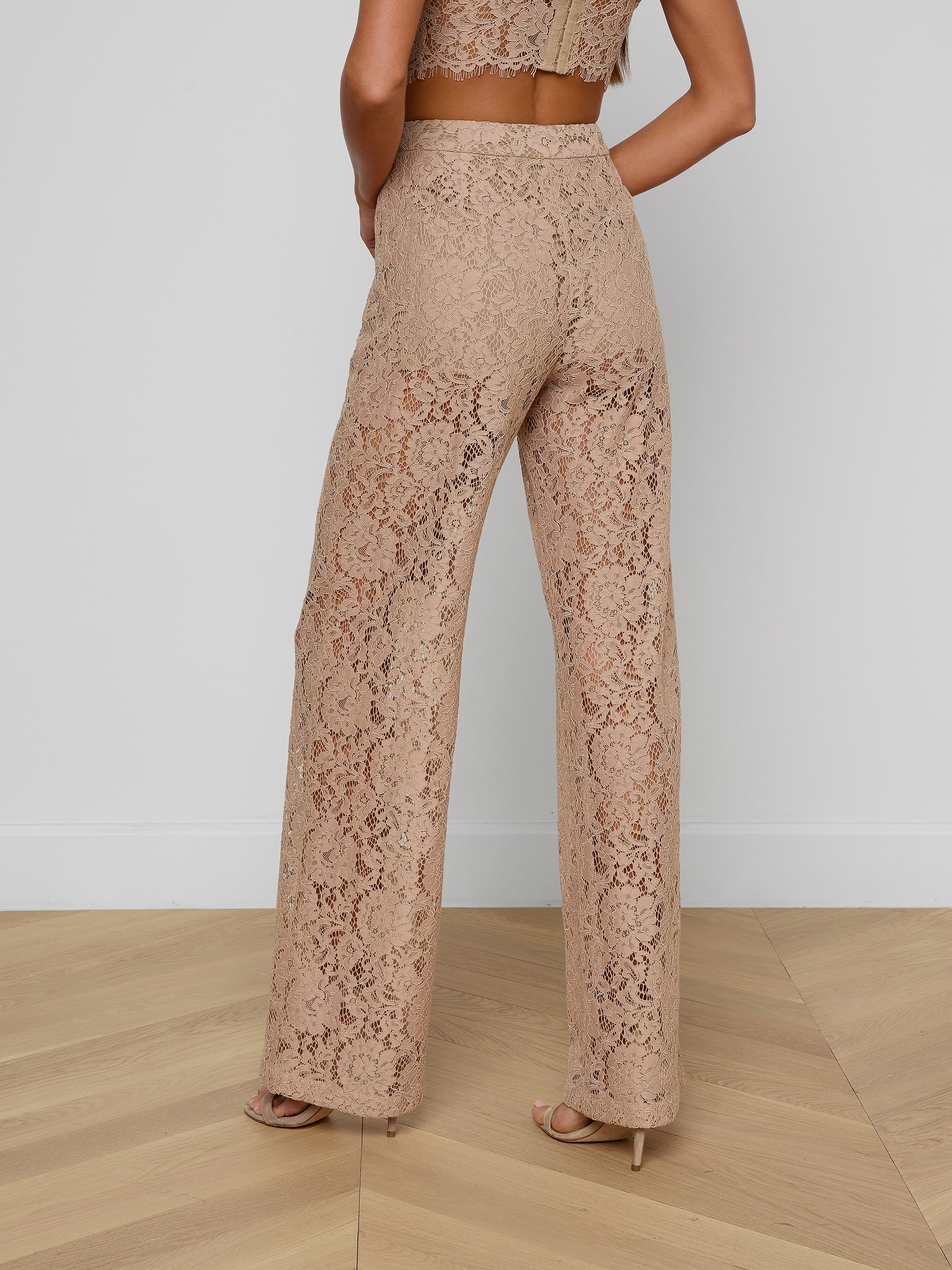 A woman is shown from the back against a plain white background, wearing the Livvy Lace Trouser—a pair of high-rise, wide-leg, tan floral lace pants—paired with heels.