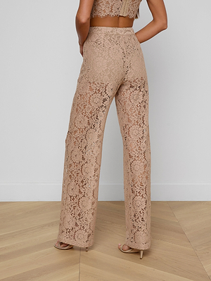A woman stands on a wooden floor wearing the Livvy Lace Trouser in cappuccino, styled with a matching lace crop top. The high-rise, wide-leg pants are semi-sheer, revealing their lining. She is photographed from the back.