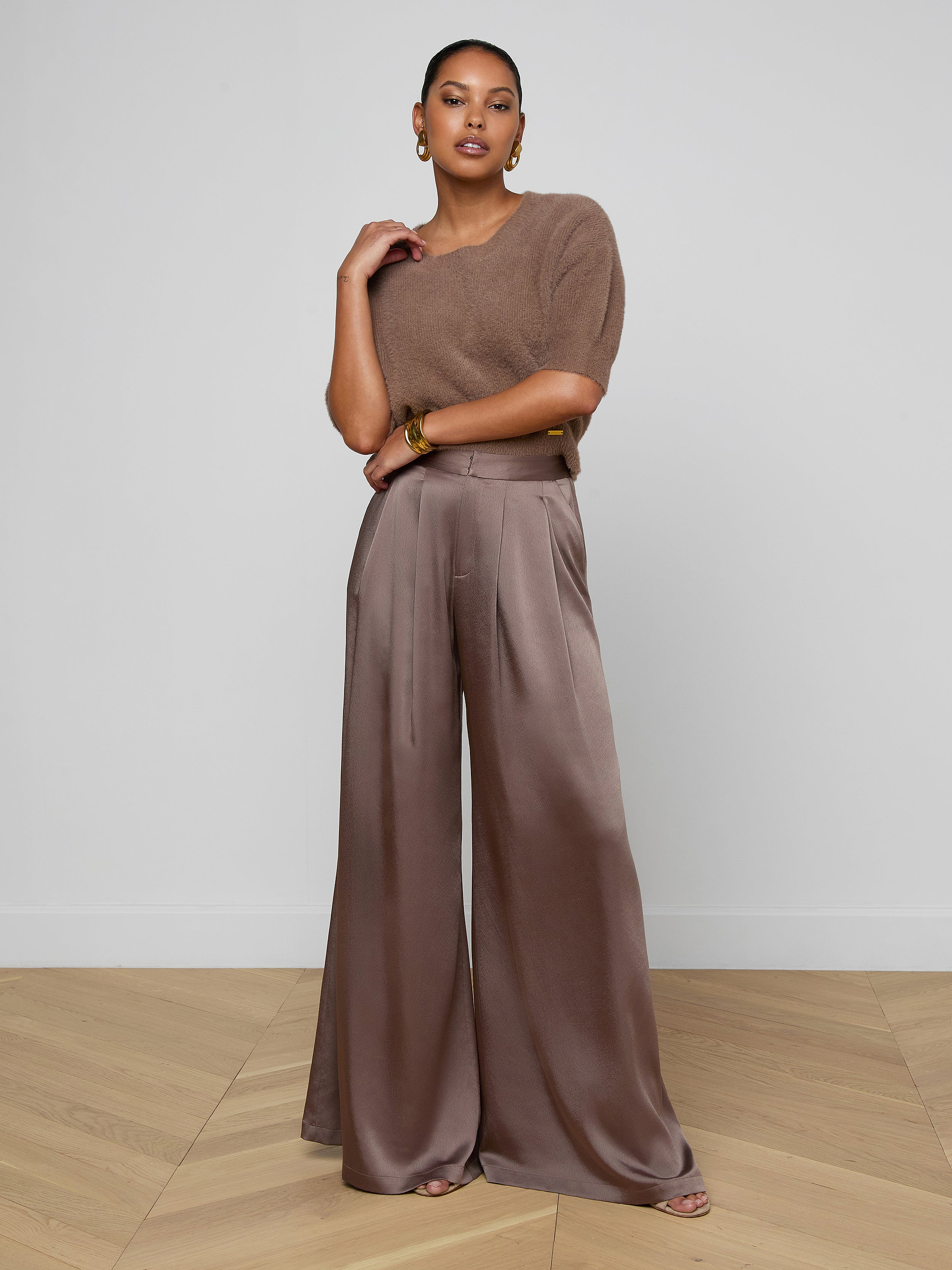 A woman stands against an off-white background, wearing a cardamom brown short-sleeve knit top paired with the Aubrie Satin Pant—a high-waisted, wide-leg trouser in a matching shade—styled with large earrings and open-toe shoes.