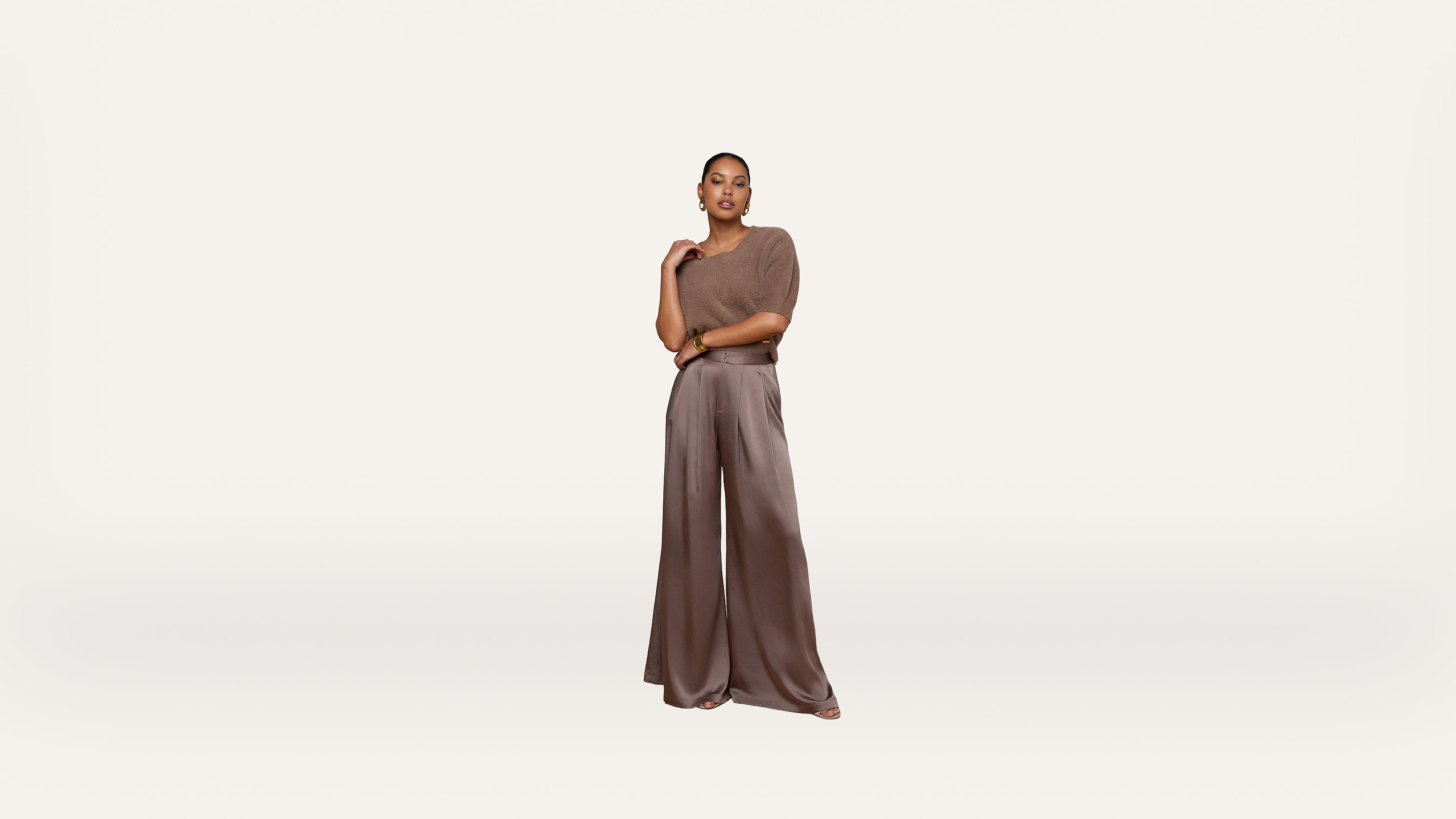 A woman stands confidently in wide-legged brown trousers and a taupe top against a plain white background.
