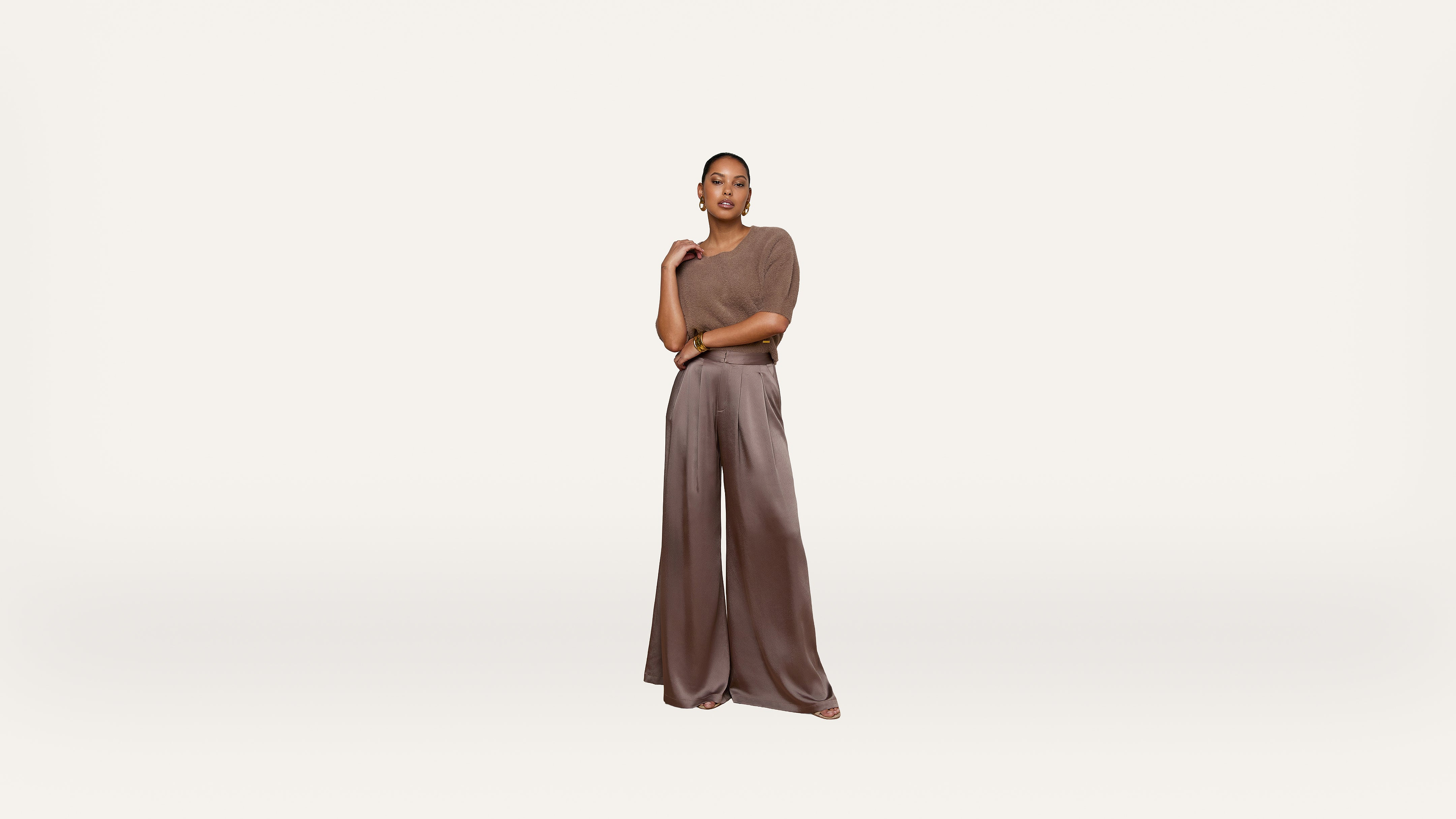 A woman stands confidently in wide-legged brown trousers and a taupe top against a plain white background.