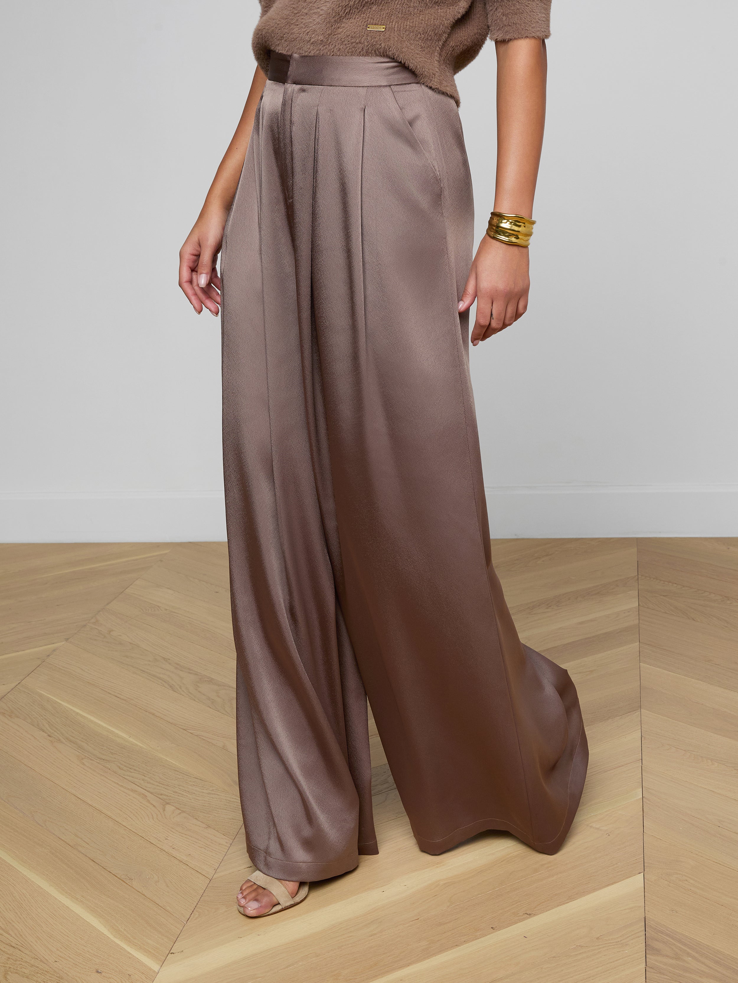 Wearing the Aubrie Satin Pant in taupe, a person pairs it with a cardamom brown knit top, gold bracelets, and sandals while posing against a plain light background.