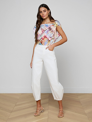 A woman stands indoors on a wooden floor, wearing a colorful top, beige heeled sandals, and the Krista Tapered Jean in white. With one hand in her pocket, she looks confidently at the camera.