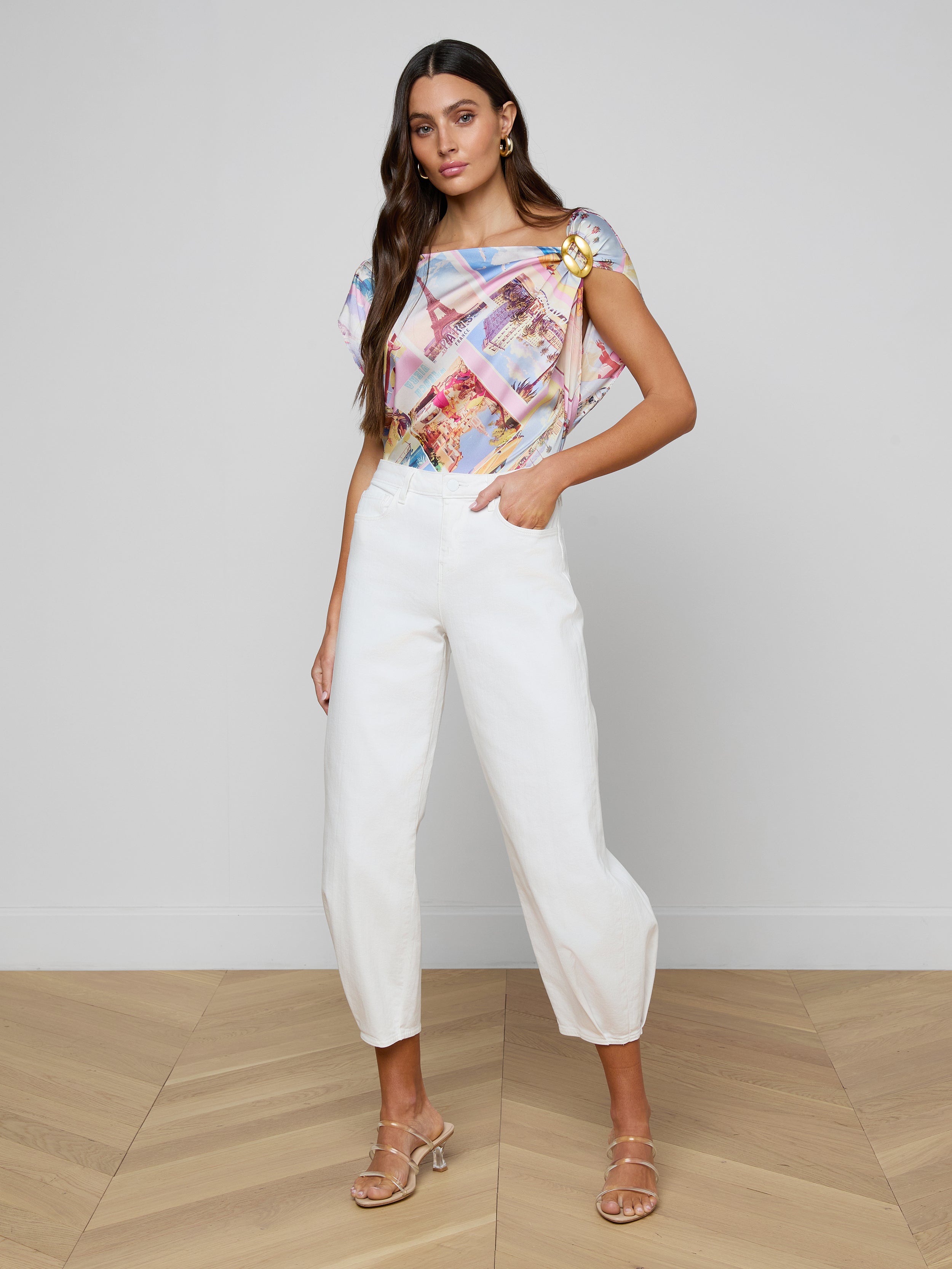 A woman in a colorful patterned short-sleeve top and Krista Tapered Jeans, paired with heeled sandals, stands against a plain light background with one hand in her pocket.
