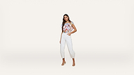 Woman poses against a plain light background, wearing a colorful off-shoulder top, white high-waisted pants, and strappy heels. She stands with one hand in her pocket, looking at the camera.