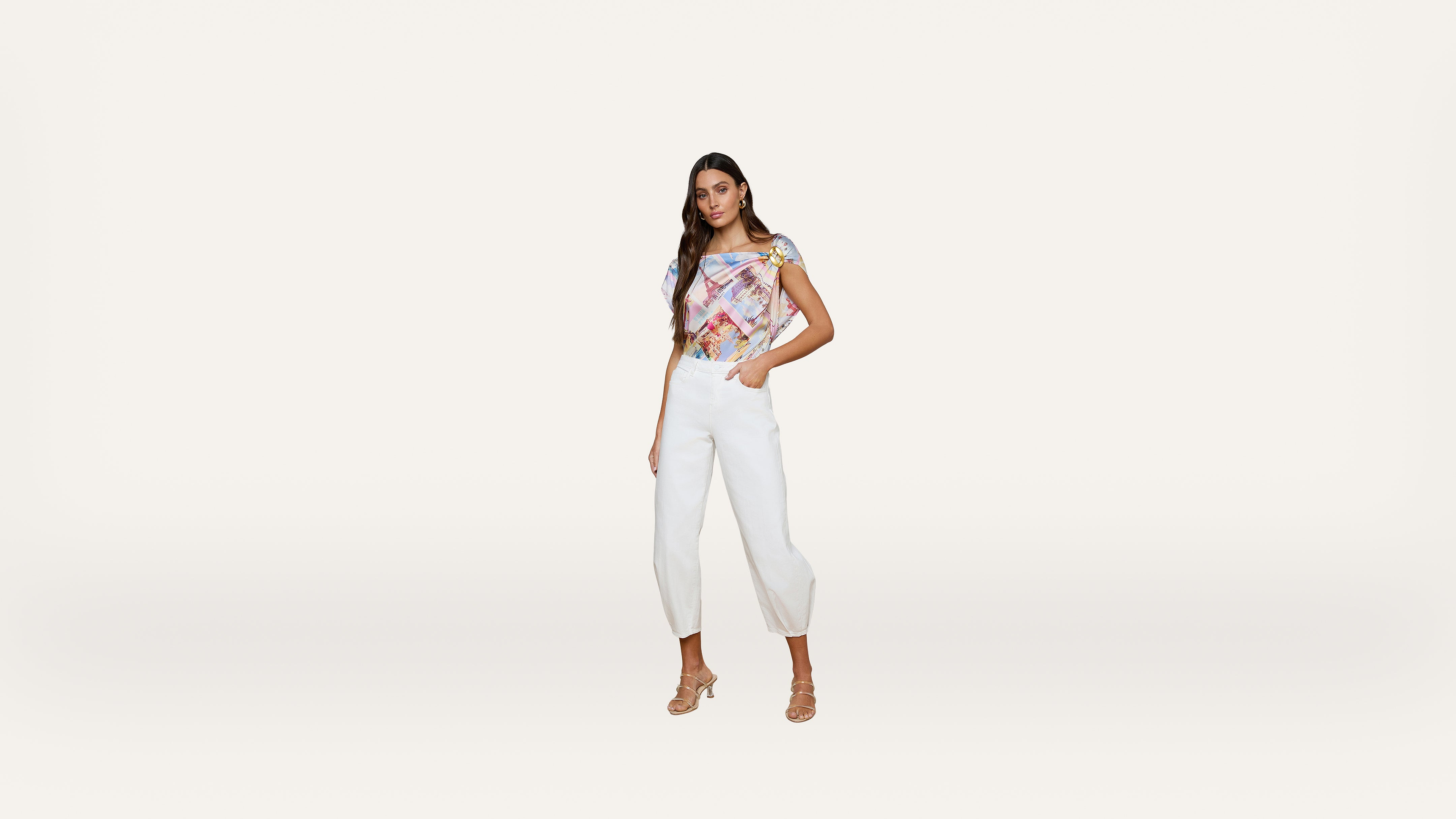 Woman poses against a plain light background, wearing a colorful off-shoulder top, white high-waisted pants, and strappy heels. She stands with one hand in her pocket, looking at the camera.
