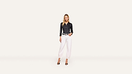 A woman stands against a white background, wearing a black long-sleeve shirt, white pants, black belt, and high-heeled shoes. With one hand in her pocket, she looks confidently at the camera.