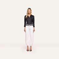 Woman stands against a plain background in a black satin blouse, Krista Tapered Jean with a belt, and black heeled sandals. She has long blonde hair and a neutral expression.