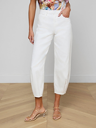A woman models the Krista Tapered Jean, a white high-rise style with tapered legs, with her left hand in the pocket. She pairs it with strappy beige heels and a colorful patterned top against a plain light-colored background.