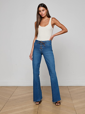 A woman poses confidently in a minimalist room wearing L'AGENCE's Charis Lace-Up Jean—high-waisted blue flare-leg jeans with a lace-up front—and a white sleeveless top, one hand on her hip and the other relaxed by her side.