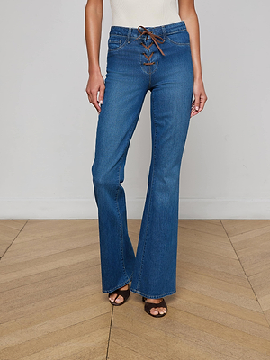 A woman stands on a wooden floor wearing the L'AGENCE Charis Lace-Up Jean—high-waisted blue flare-leg jeans with a brown lace-up front—paired with black open-toe heels; her white top is partially visible.