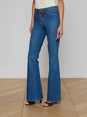 A person wears the L'AGENCE Charis Lace-Up Jean—high-waisted, blue stretch denim with a brown lace-up detail—paired with brown open-toe heels, standing on a wooden floor against a plain white wall.