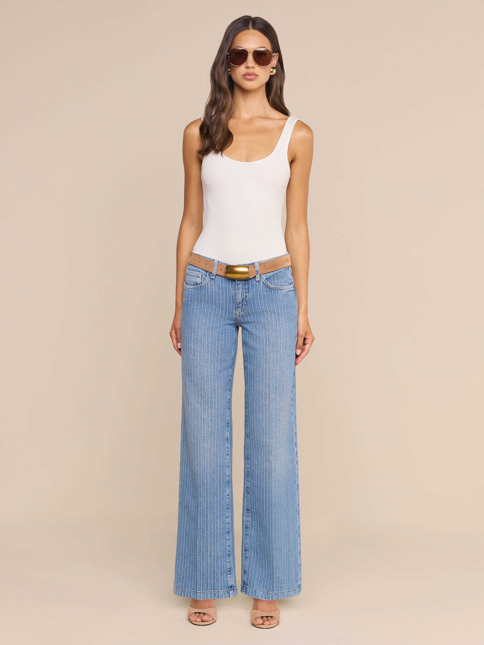 Eine Frau blickt vor einem beigen Hintergrund nach vorne; sie trägt eine Sonnenbrille, ein weißes, ärmelloses Oberteil, die „Joanne Low-Rise Wide-Leg Jean“ mit einem braunen Gürtel sowie beigefarbene Sandalen mit Absatz.