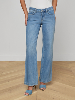 Wearing Joanne Low-Rise Wide-Leg Jeans in pinstriped indigo, a white top, and blue open-toe heels, a person stands on a wooden floor with hands near pockets; the head is not visible.