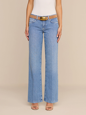 Une personne porte un jean « Joanne » taille basse à jambes larges de couleur indigo, avec une ceinture beige et un haut blanc, debout devant un fond neutre.