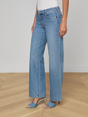 A woman stands on a wooden floor wearing Joanne Low-Rise Wide-Leg Jeans in pinstriped indigo, paired with a white top and blue heeled sandals. Her face is not visible.