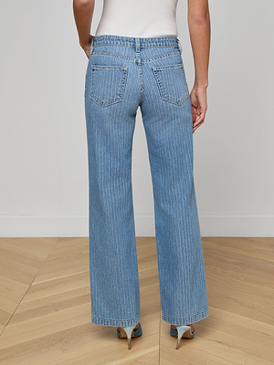 Someone stands on a light wood floor, facing away, wearing the Joanne Low-Rise Wide-Leg Jean in light blue with subtle indigo pinstripes, two back pockets, and heeled sandals.