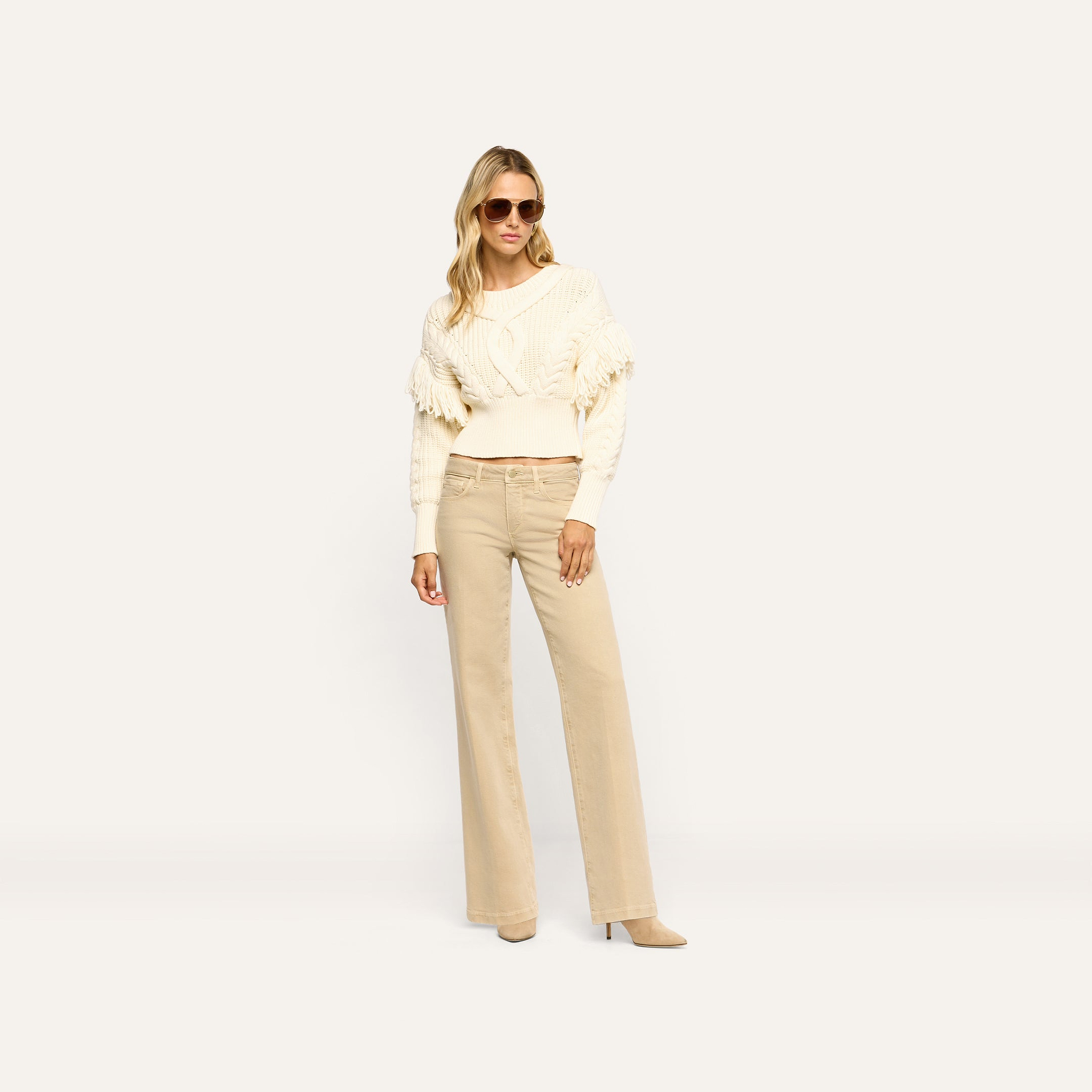 A woman wearing a cream cable-knit sweater with fringe detailing and tan Joanne Low-Rise Wide-Leg Jean.