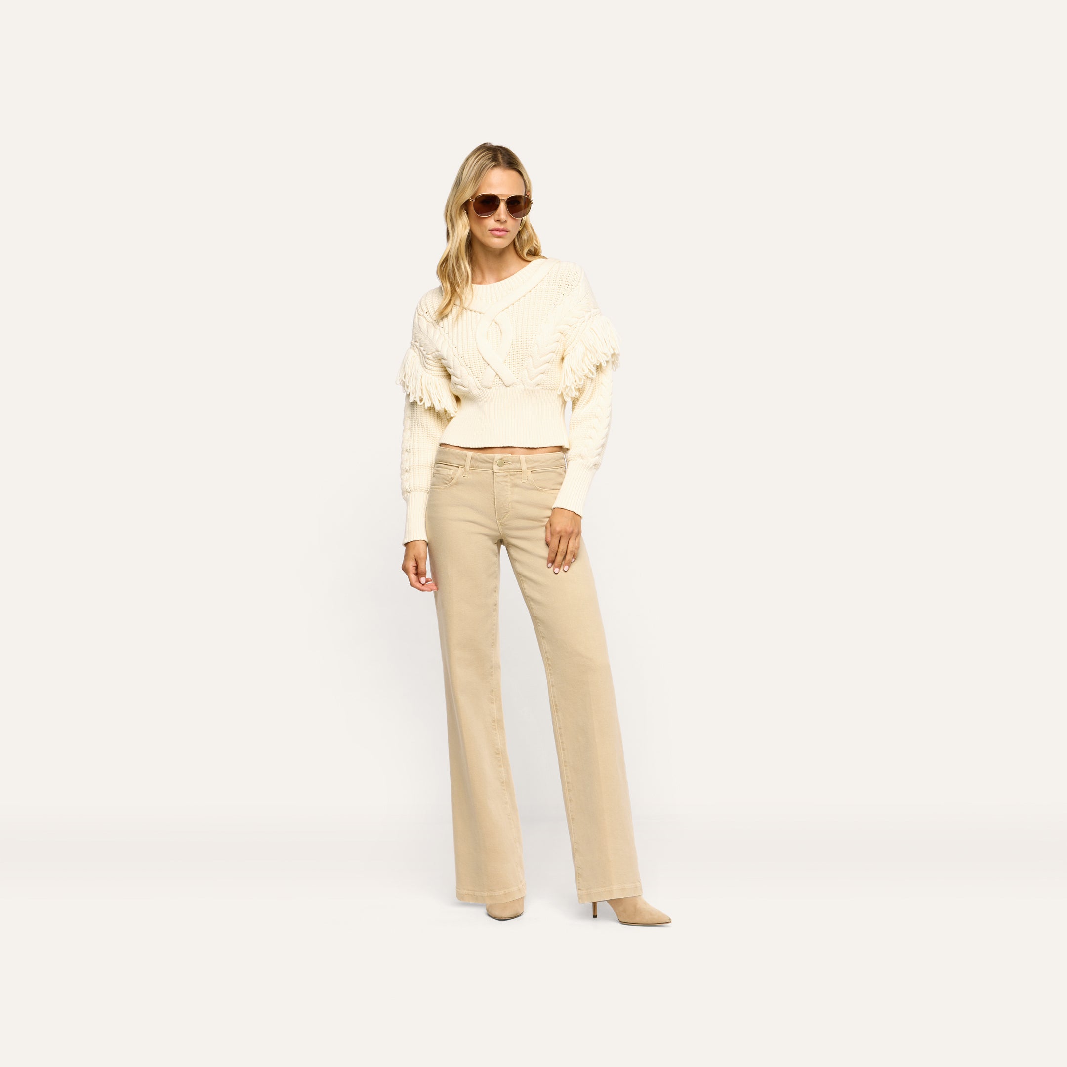 A woman wearing a cream cable-knit sweater with fringe detailing and tan Joanne Low-Rise Wide-Leg Jean.