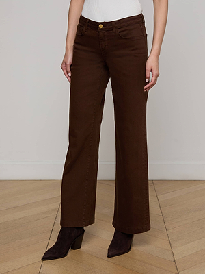 A person wearing L'AGENCE Joanne Low-Rise Wide-Leg Jeans in brown stretch denim, a white top, and dark brown heeled boots stands on a light wooden floor against a plain white wall. Their head is not visible in the image.