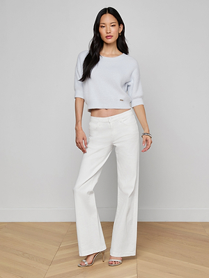 A woman with long dark hair stands indoors by a plain light gray wall, wearing a cropped sweater, heeled sandals, and the Joanne Low-Rise Wide-Leg Jean in white. She poses relaxedly with one hand on her hip.