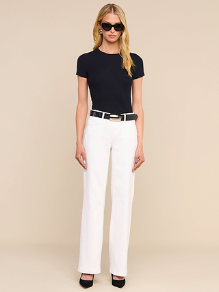 Donna dai lunghi capelli biondi che indossa occhiali da sole neri, una maglietta nera a maniche corte, una cintura nera, jeans Joanne a vita bassa e gamba larga con lavaggio bianco e tacchi neri, in piedi su uno sfondo beige.