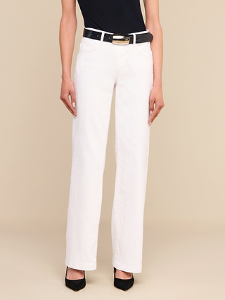 Une personne portant un jean « Joanne » taille basse à jambes larges de couleur blanche, assorti dune ceinture noire à boucle dorée, dun haut noir et descarpins noirs, se tenant debout devant un fond beige.