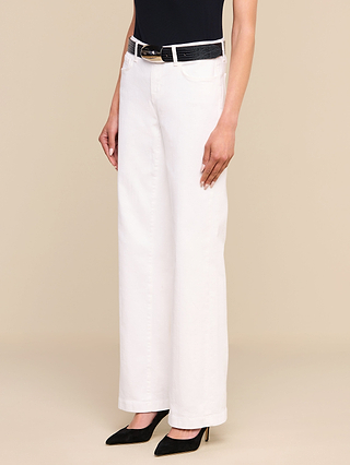 Une personne portant un jean « Joanne Low-Rise Wide-Leg » blanc, assorti dune ceinture noire, dun haut sans manches noir et descarpins noirs à bout pointu, se tient debout devant un fond beige.