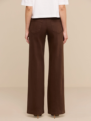 Una persona está de espaldas, mostrando únicamente la parte baja de la espalda y las piernas, vestida con unos vaqueros de talle bajo y pierna ancha de la marca Joanne, combinados con una camiseta blanca de manga corta, sobre un fondo beige.