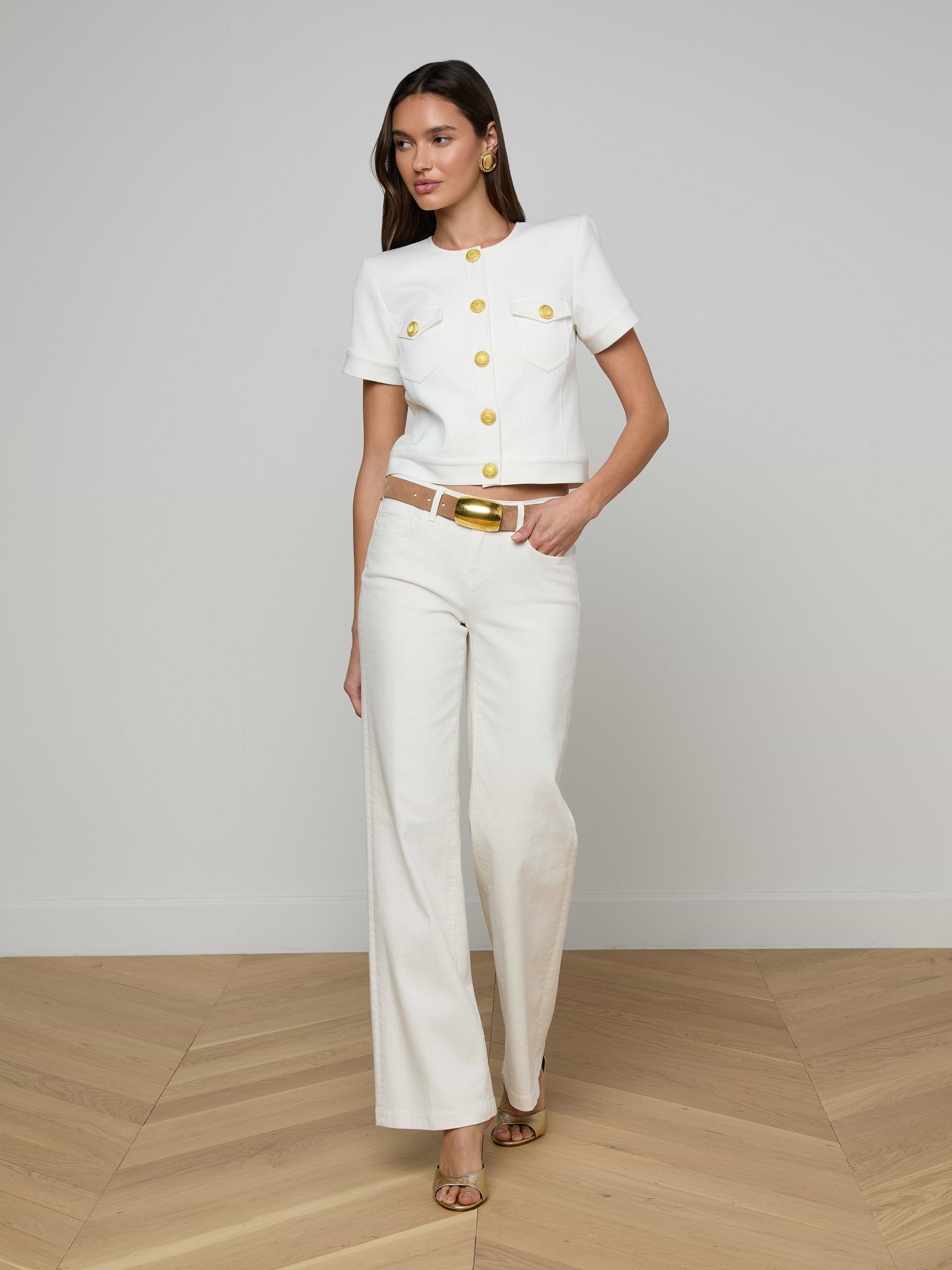 A woman stands in a minimalist room wearing Joanne Low-Rise Wide-Leg Jeans in ecru wash, paired with a white short-sleeve jacket with gold buttons, a white belt featuring a gold buckle, and open-toe heels.