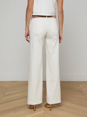 A person stands facing away on a light wood floor, wearing Joanne Low-Rise Wide-Leg Jeans in an ecru wash with beige heeled sandals, set against a plain white wall.