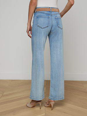 A woman stands on a wooden floor with her back to the camera, wearing Joanne Low-Rise Wide-Leg Jeans in a light wash, paired with heeled sandals and a white sleeveless top. The jeans feature back pockets and a sheer waistband.
