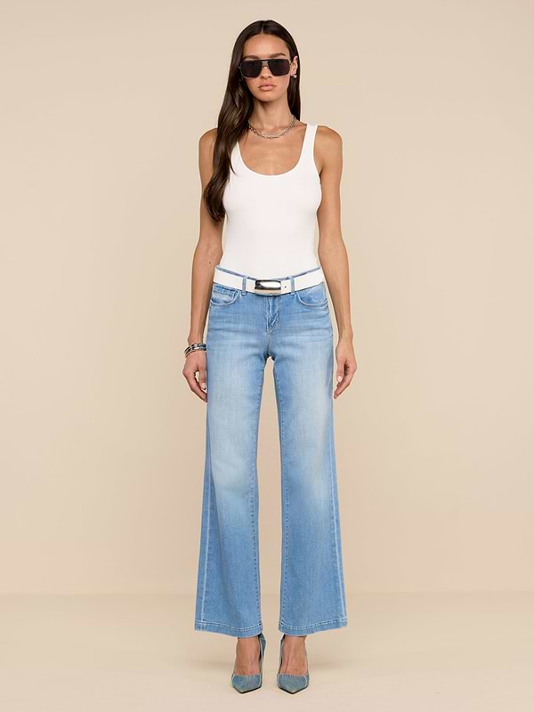 Une femme se tient sur un fond beige et porte des lunettes de soleil, un haut blanc sans manches, le jean Joanne Low-Rise Wide-Leg en délavage indigo clair, une ceinture blanche, des talons bleus et des bracelets argentés.