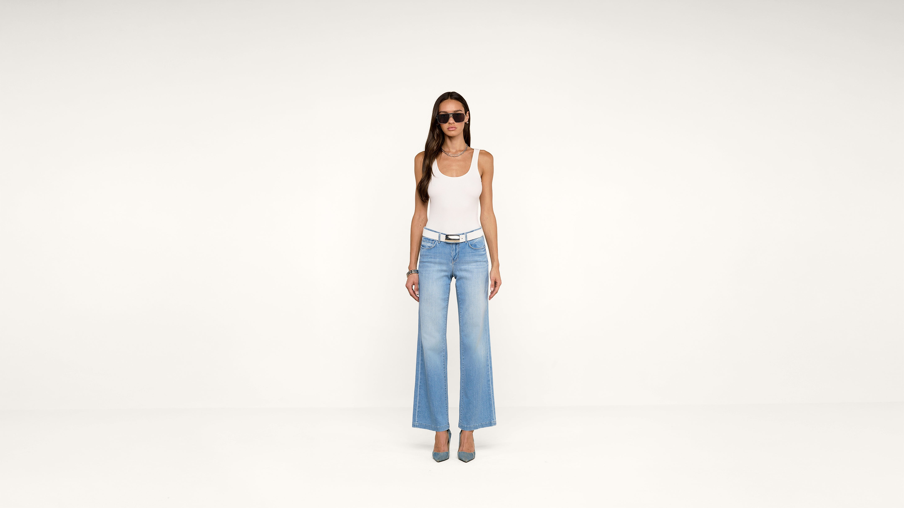 A woman in sunglasses, a white tank top, and the Dee Capri Pant in blue stands in heels against a plain white background.