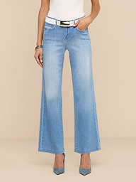 Une personne vêtue dun jean large à taille basse Joanne délavage indigo clair, dune ceinture blanche, dun haut blanc, dun bracelet et de chaussures bleues à bout pointu se tient debout sur un fond beige uni.