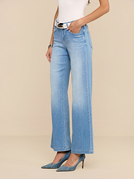 Une personne porte un jean Joanne à taille basse et à jambe large en délavage indigo clair, associé à une ceinture blanche, un haut sans manches blanc et des talons bleus à bouts pointus, sur un fond beige uni.